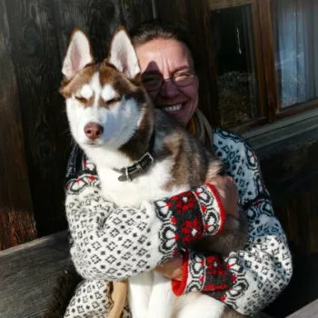 Auf einer Bank in einem bunten Norwegerpullover sitzt vor einer Holzhütte eine lachende Frau mit auf dem arm einen jungen sibirischen Husky der die Augen zukneift.