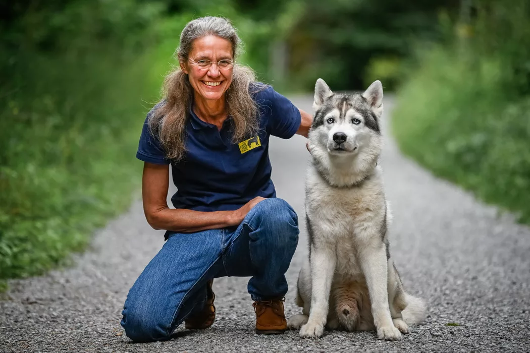 Die Hundetrainerin schaut lachend in die Kamera, trägt ein blaues Poloshirt und eine Jeans mit braunen Wildleder Schuhen. Daneben sitzt aufrecht und ernst schauend ein grauer sibirischer Husky Rüde